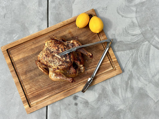 Roast chicken resting on a wooden board with a kitchen thong and a pair of lemons on the side