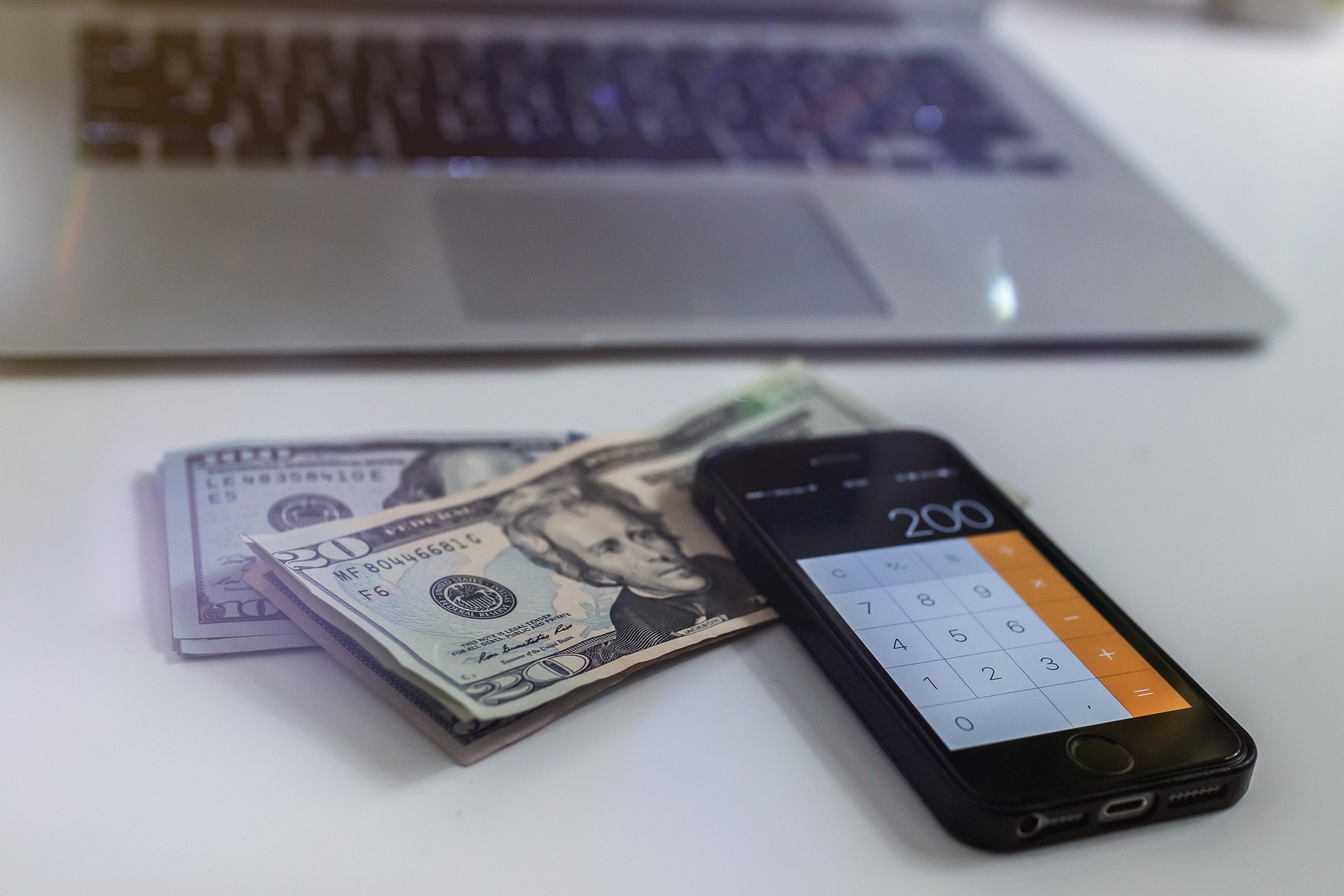 Cellphone with the calculator app visible, while it is is placed on top of dollar bills and beside a laptop